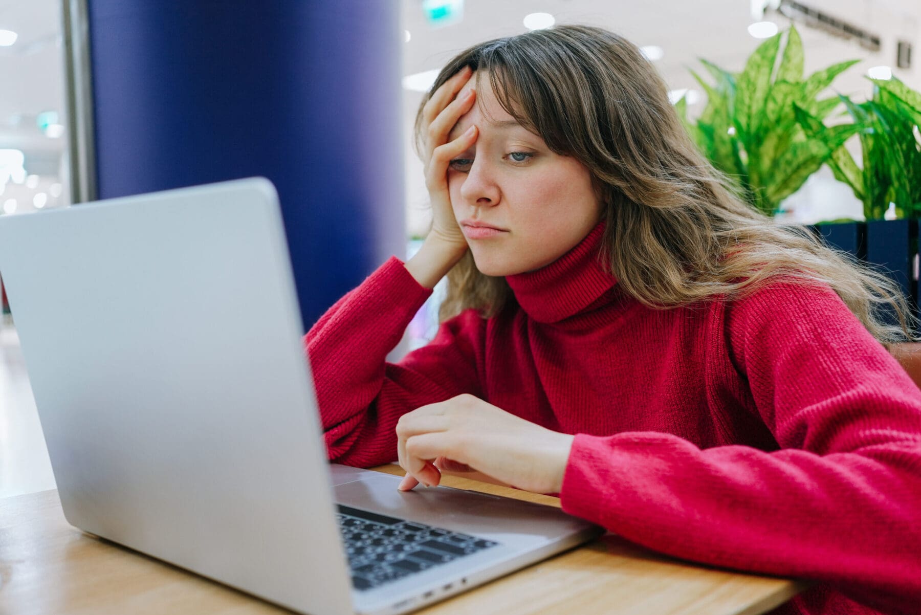 Young woman wearing red turtleneck sweater is working on a laptop at a table in a cafe, looking tired and supporting her head with one hand while using the trackpad with the other