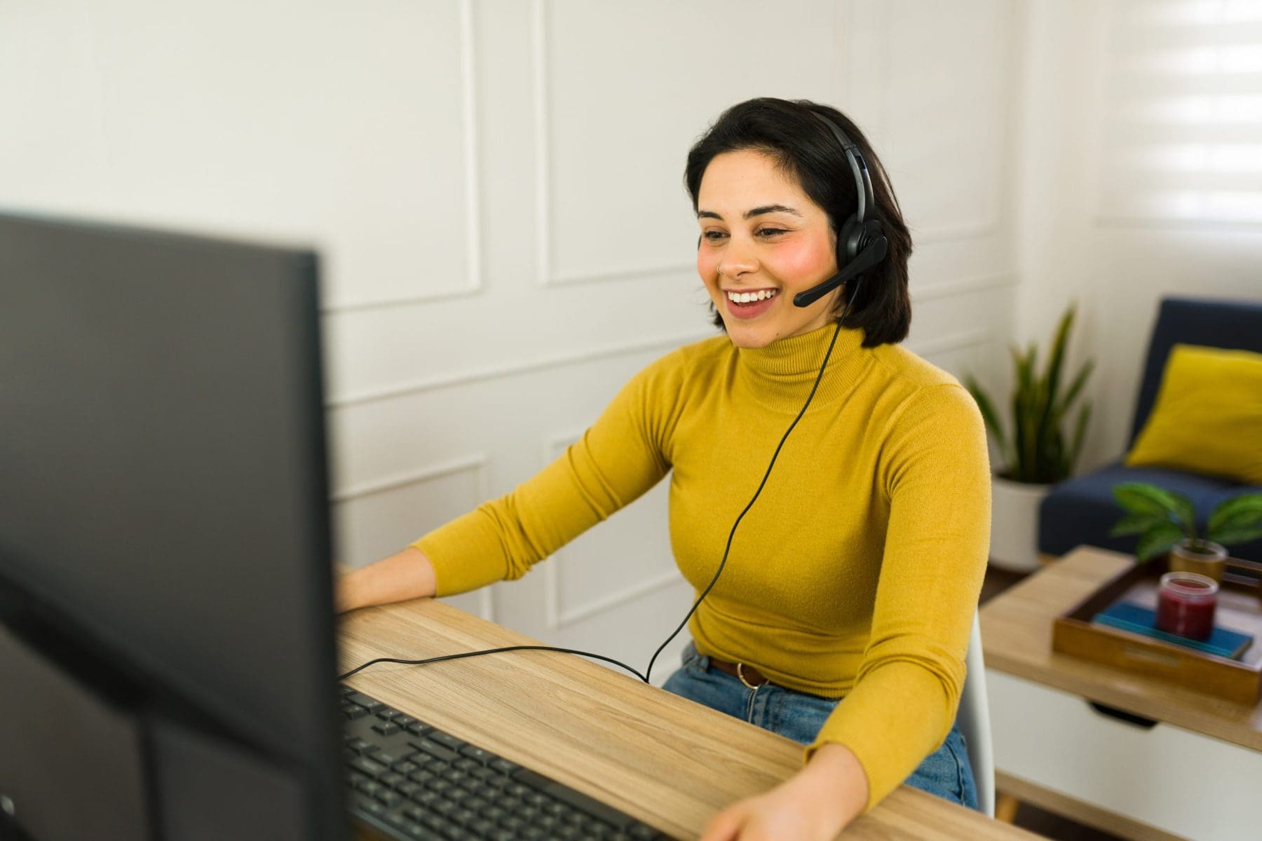 A woman wearing a headset is smiling while working at a computer in a bright, home office setting.