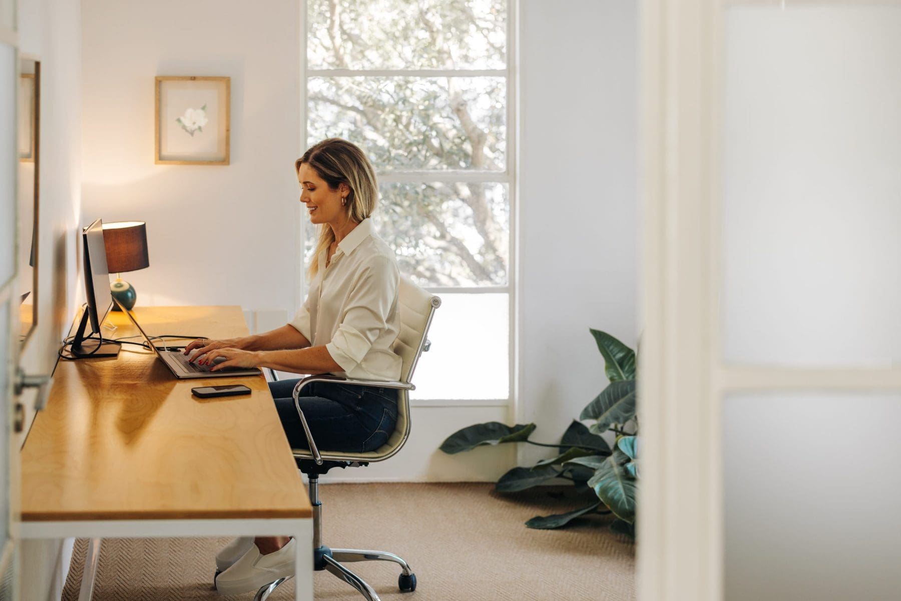 A woman types on a laptop at a long desk in a bright room with large windows and indoor plants.