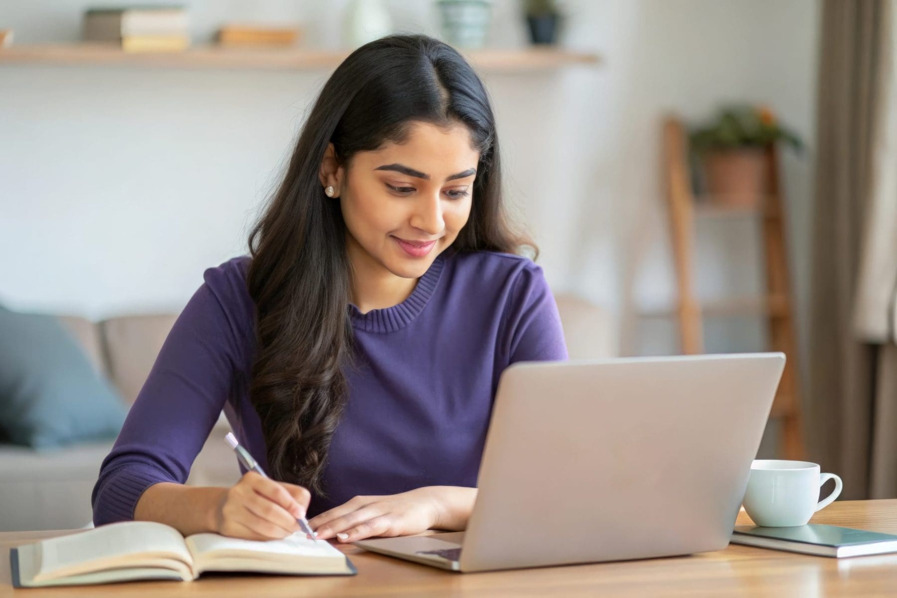 A woman writes in a notebook while looking at her laptop, working from a comfortable home environment.