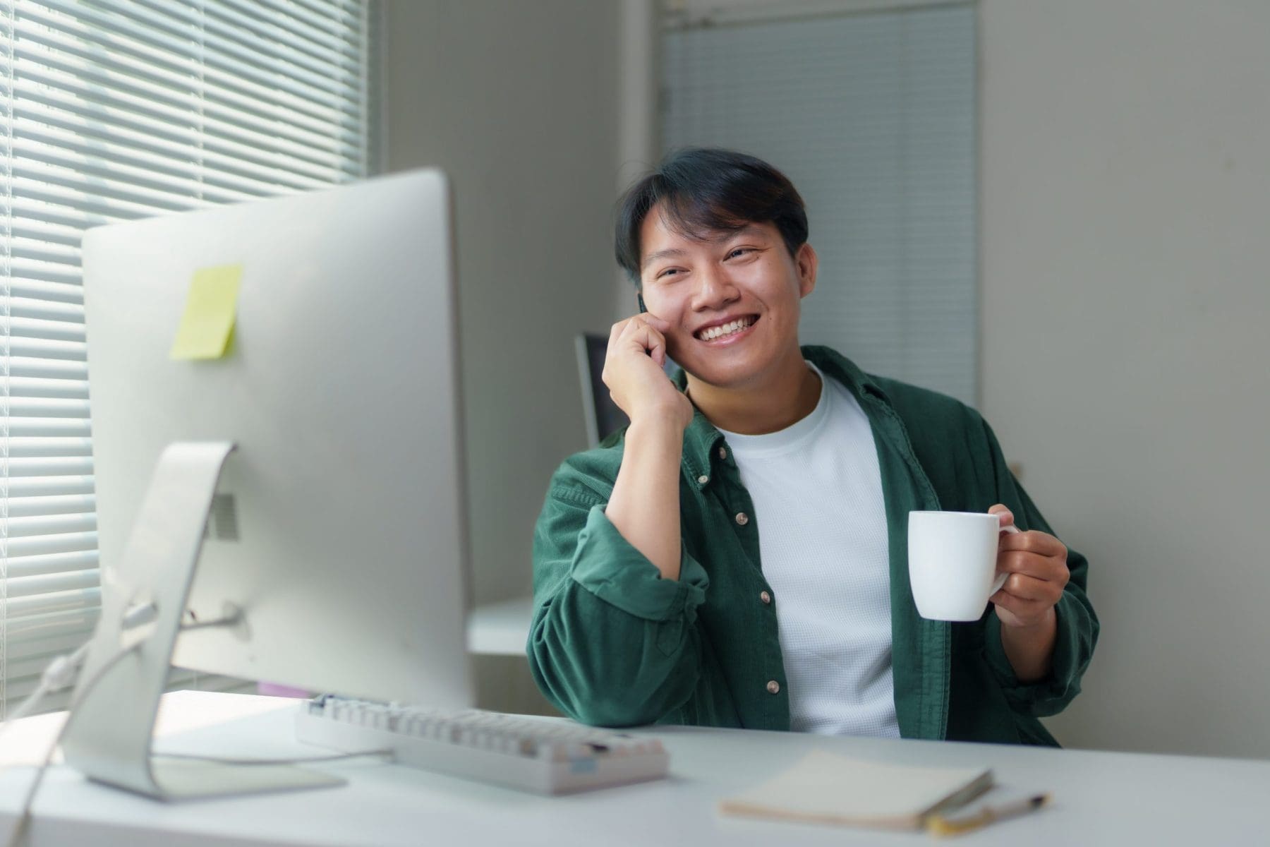 Young man working remotely at a desk, using a desktop computer, chatting on the phone, and enjoying a cup of coffee