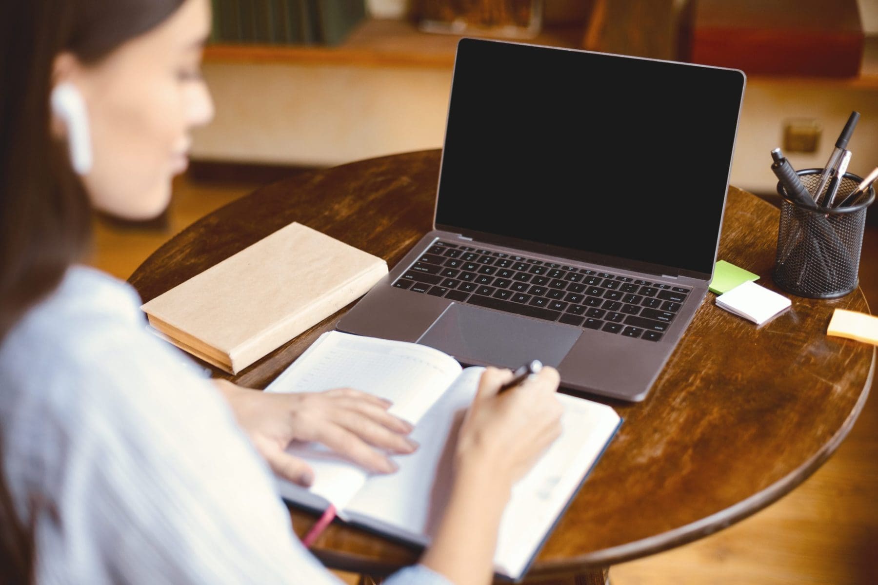 Young woman wearing earbuds sitting at table and writing in her notebook, taking notes and using laptop with black blank screen..
