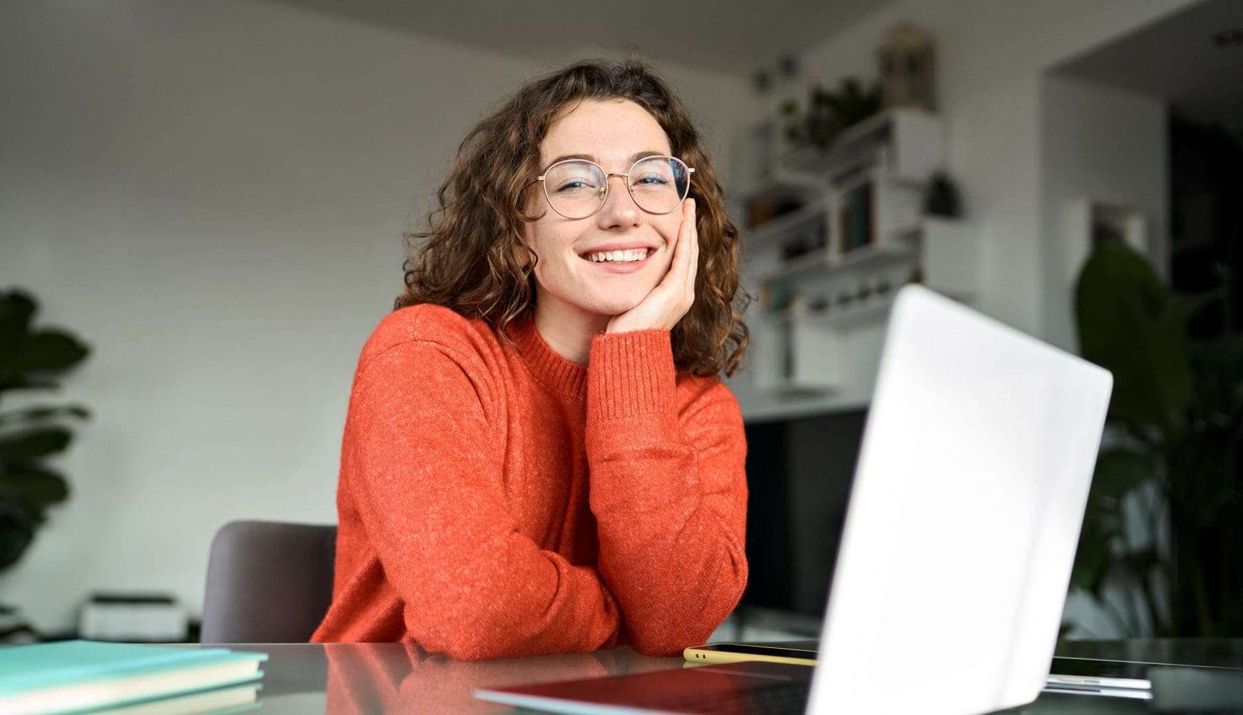 Portrait of happy young woman or student wearing glasses sitting at desk at home office with laptop computer looking at camera.