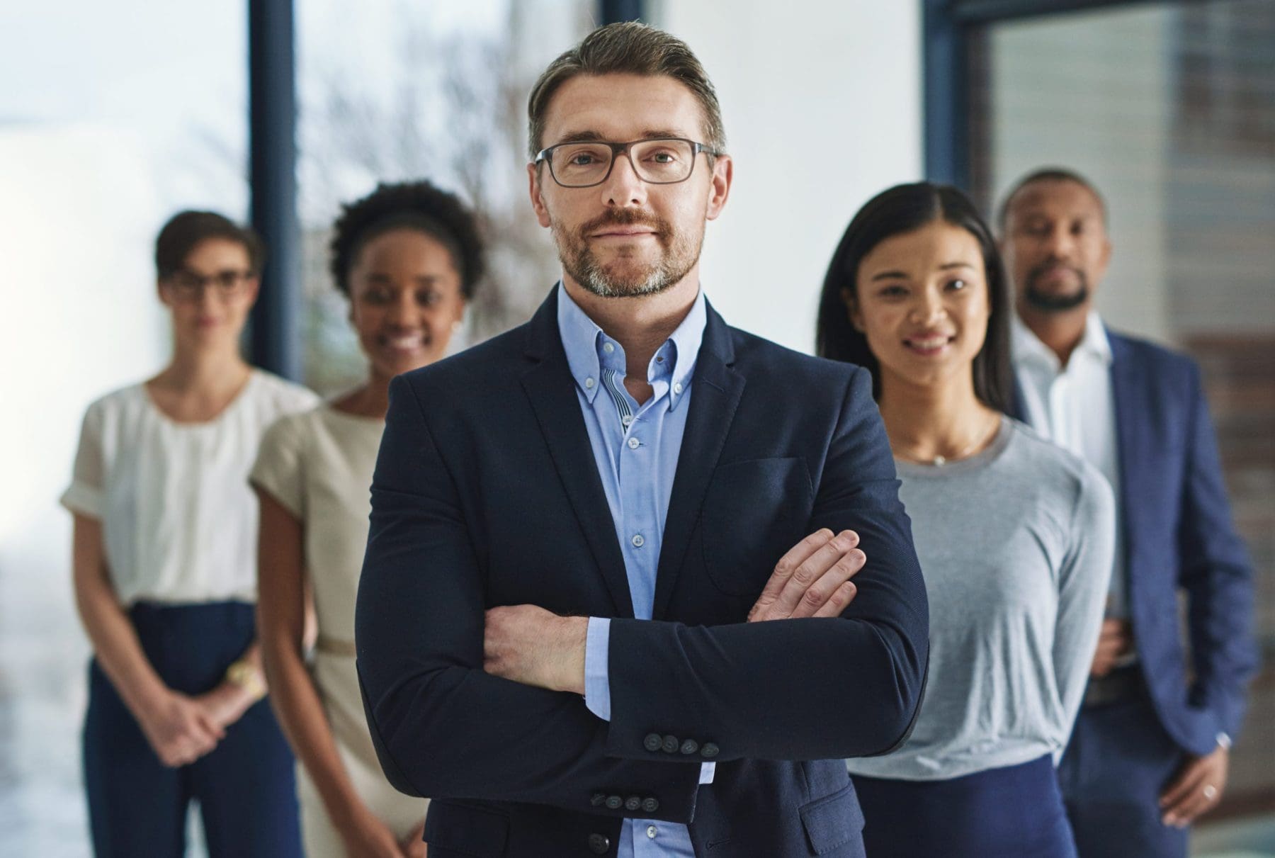 A man stands with arms crossed, slightly ahead of a diverse group of professionally dressed individuals, embodying confidence and team solidarity.