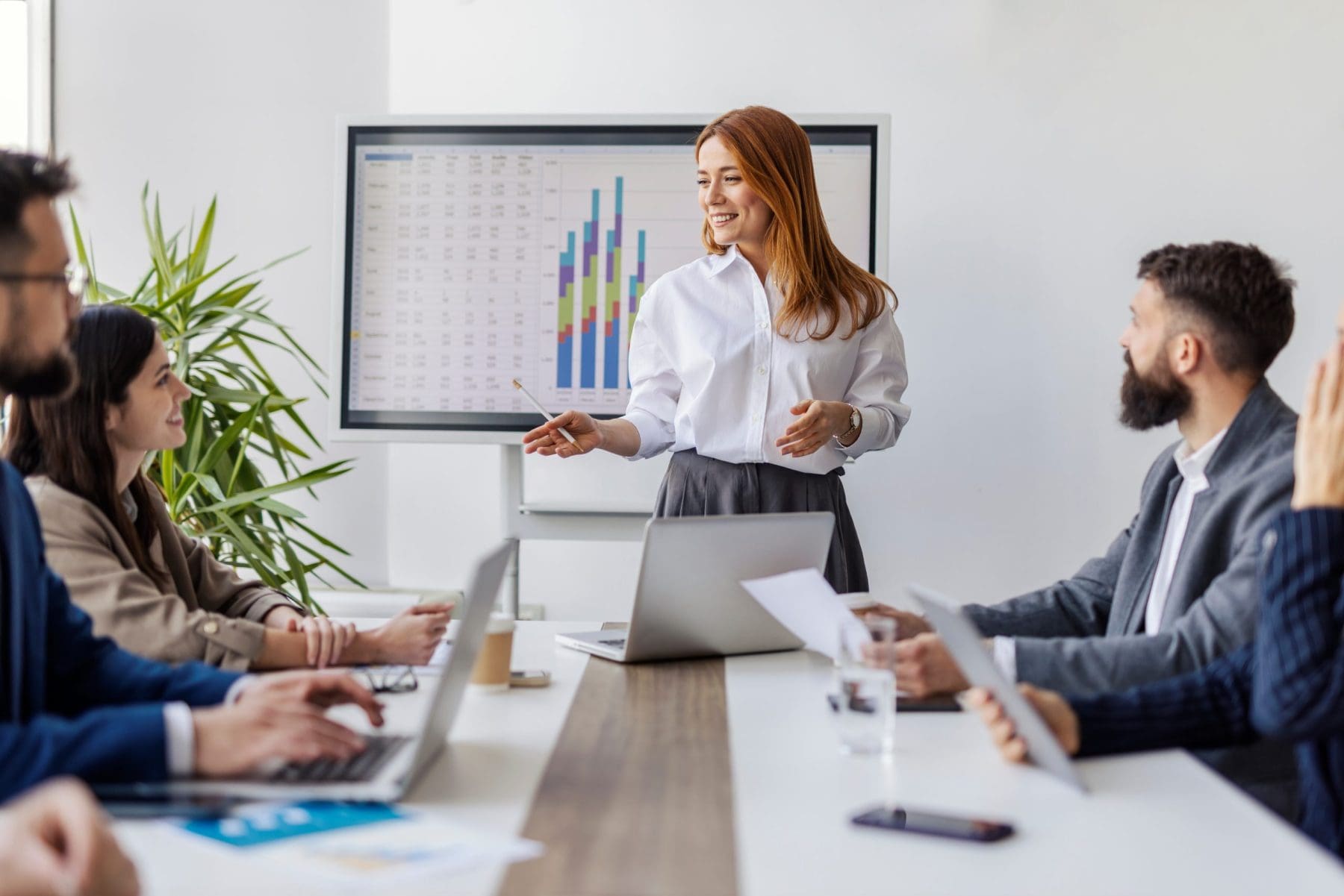 A woman leads a meeting, standing in front of a screen filled with charts. She addresses a table of attentive team members in a bright, professional environment