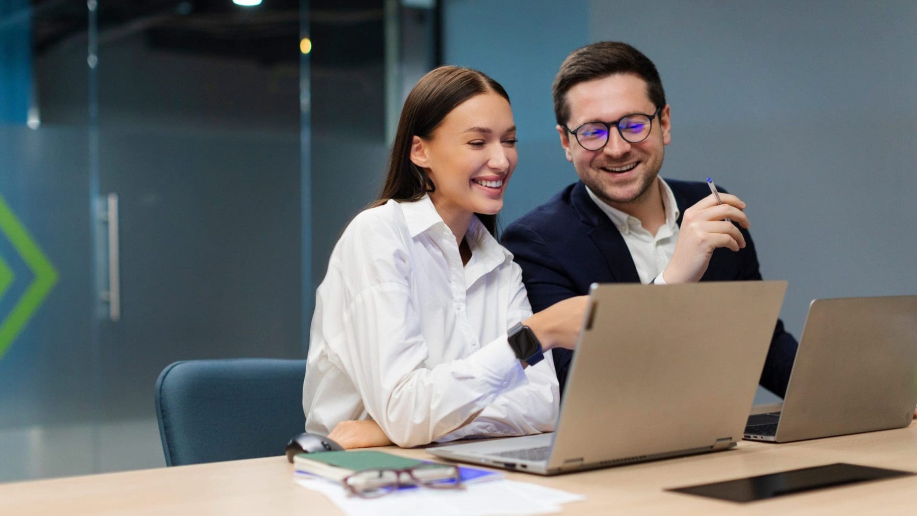 Two colleagues review a laptop together, exchanging smiles in a collaborative moment. They appear engaged and aligned as they work side by side.