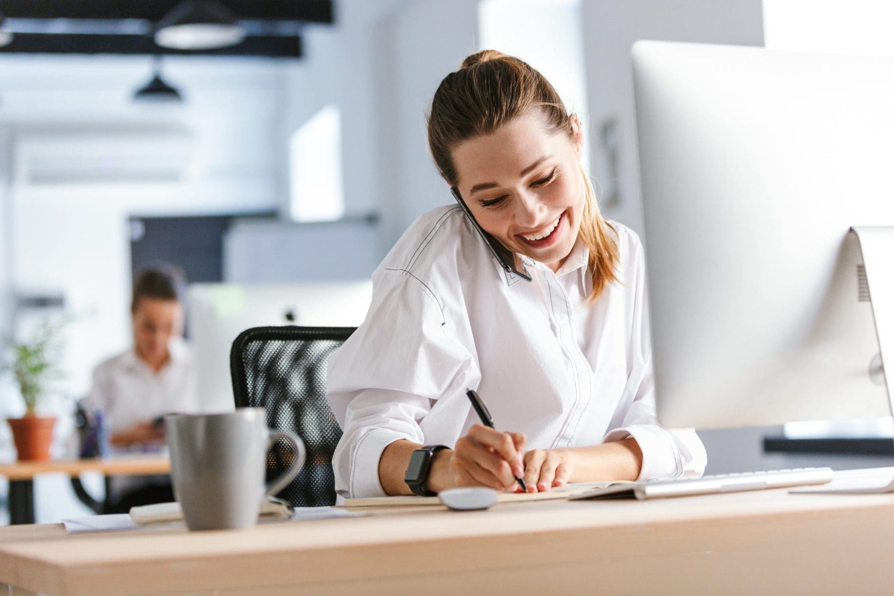 A woman balances a phone call while writing notes, radiating energy and professionalism. Her relaxed posture and smile convey confidence and efficiency.