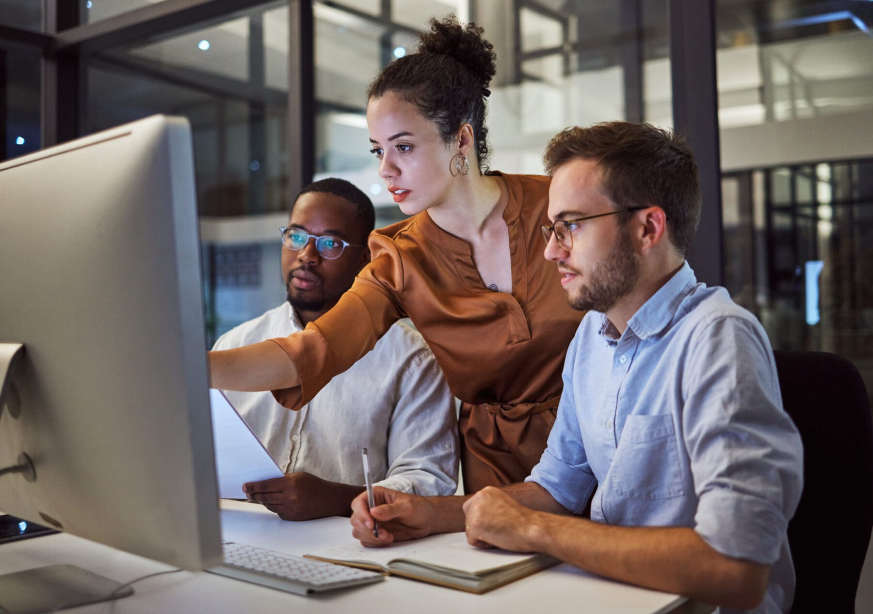 Woman points at computer and explains to two young man sitting on desks in office space.