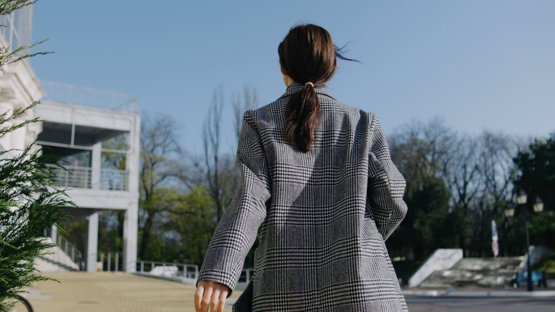 A woman in a stylish checked blazer strolls away from a contemporary building under a bright blue sky.