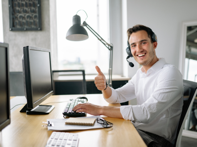 Man workin on desktop, wearing headset with thumbs up and smiling.