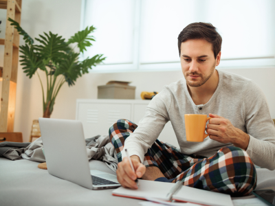 Man holding coffee cup and working on laptop while sitting in bed.