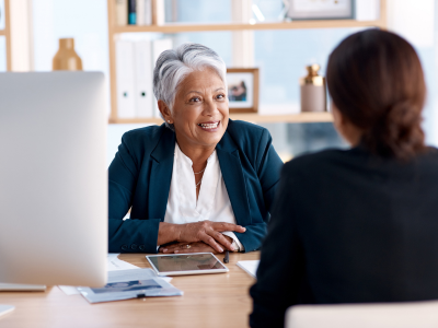 Woman sitting on desk smiling to another woman on another end of desk.